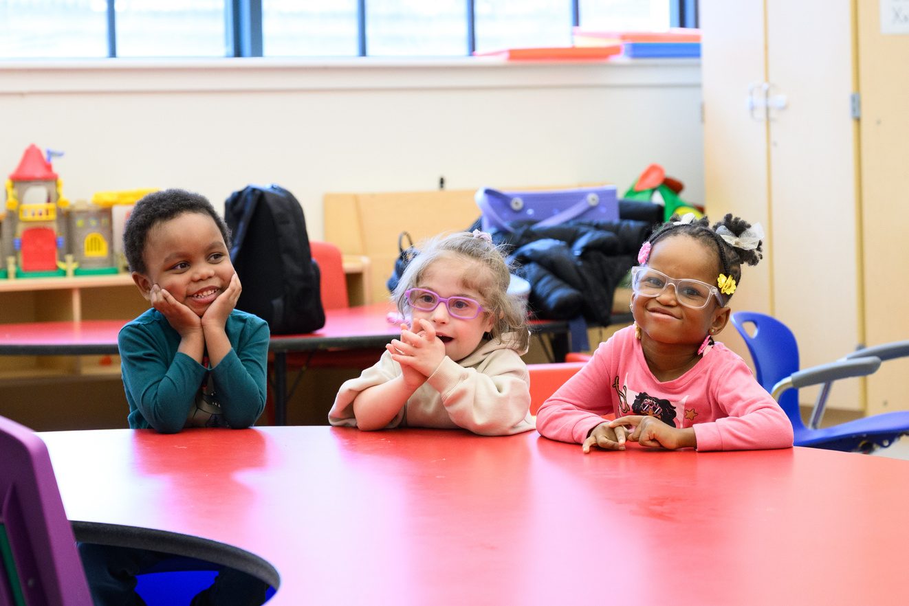 Three children sitting together at a red table on the first day of school, resting their chins on their hands and smiling at the camera.