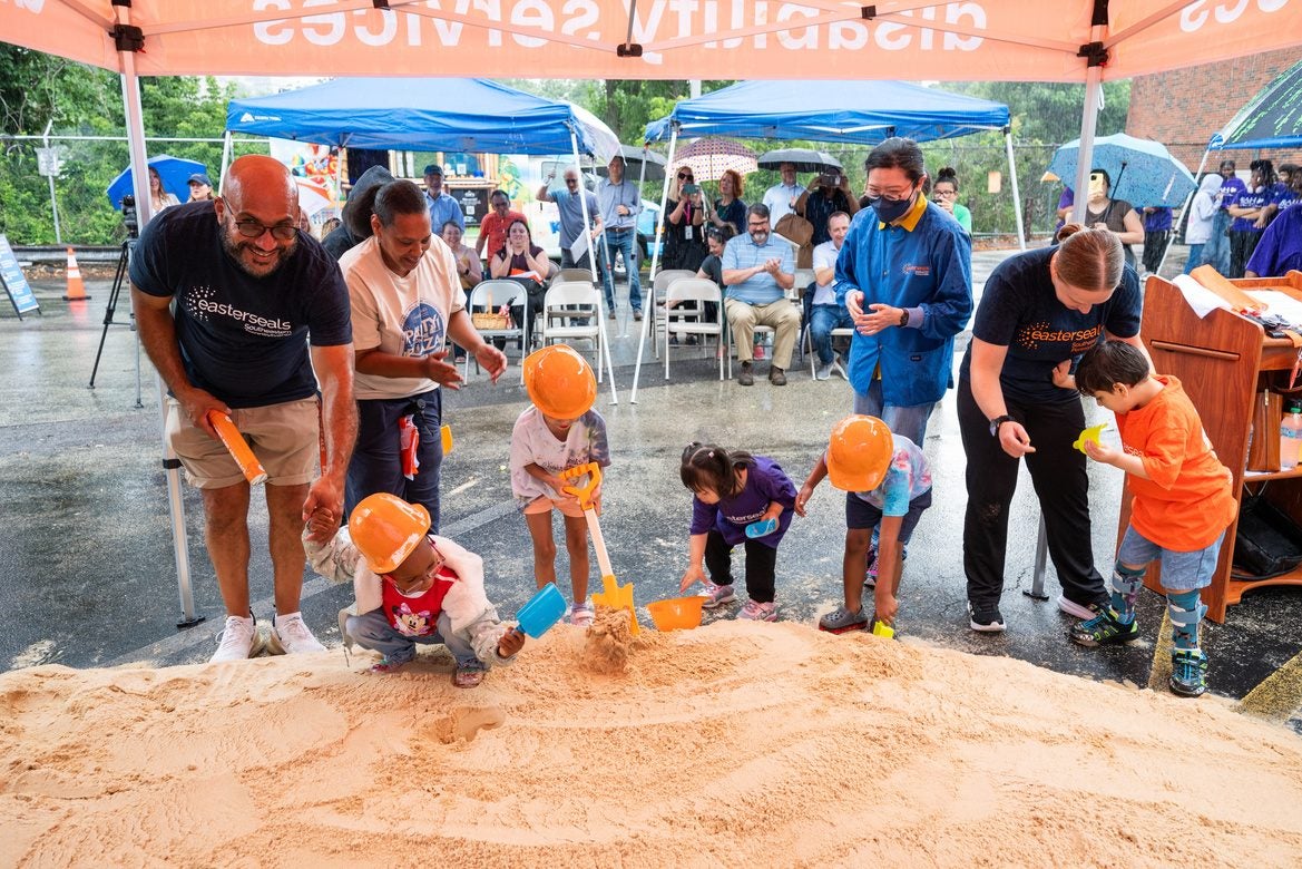 Staff, families, and children using small shovels together in a ceremonial pile of sand at the groundbreaking event.