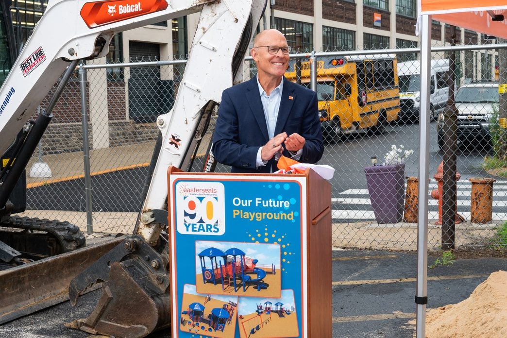 An Easterseals leader in a suit speaking at an outdoor podium labeled 'Our Future Playground,' with construction equipment visible behind him.
