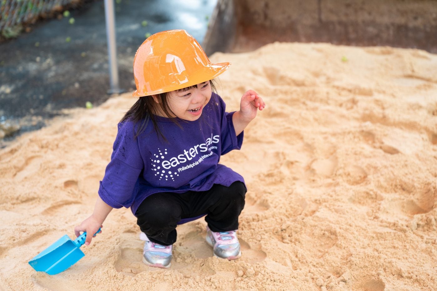 A child wearing an orange toy hard hat and an Easterseals shirt, crouched in a sand pit with a small shovel at the playground groundbreaking event.