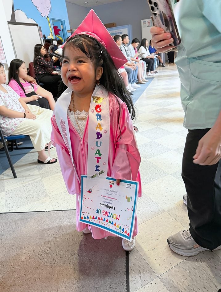 A young Easterseals graduate wearing a pink cap and gown, smiling and holding her diploma at a Moving Up ceremony while classmates and families look on.