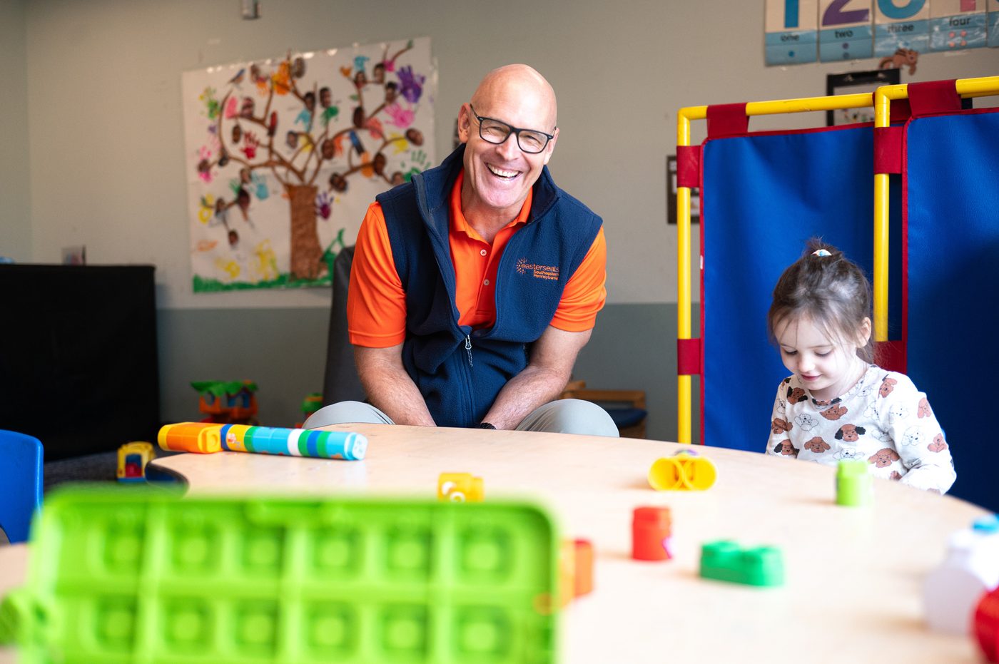 Tim, Easterseals SEPA's CEO, kneeling at a low table and smiling as a young child in a classroom plays with colorful stacking blocks.